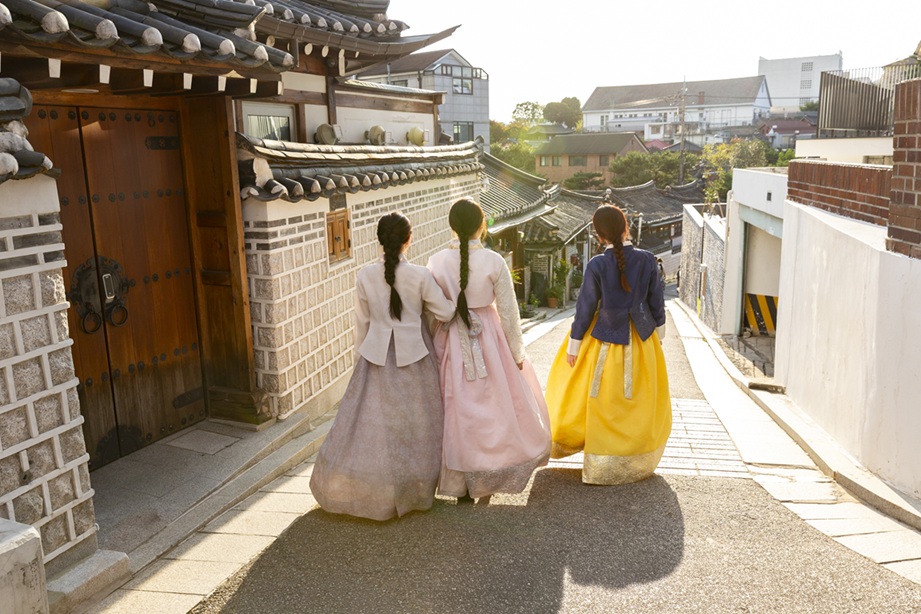 한복을 입은 세 명의 여성이 따뜻한 햇살 아래 전통적인 한옥 마을 골목길을 다정하게 걷는 모습