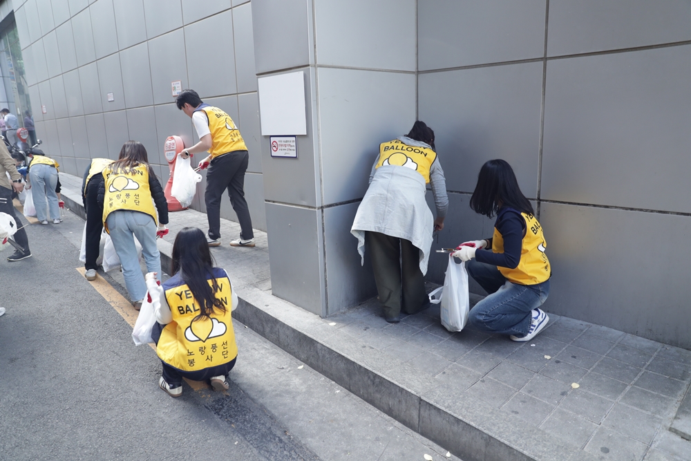 노랑풍선 봉사단 조끼를 입은 사람들이 건물 주변 거리에서 쓰레기를 줍는 환경 정화 봉사